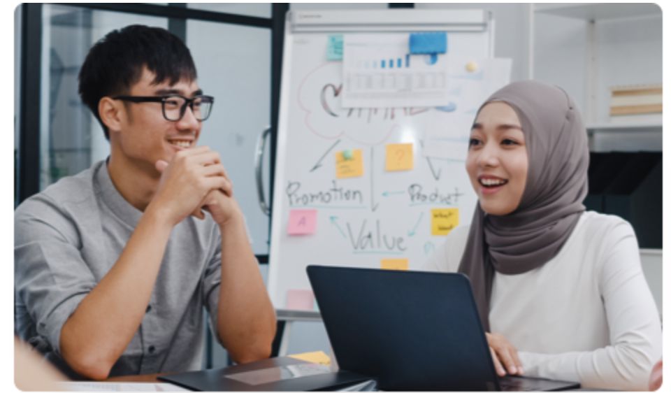 A young Muslim girl with a laptop in front of her is seated at a table with an Asian co-worker.