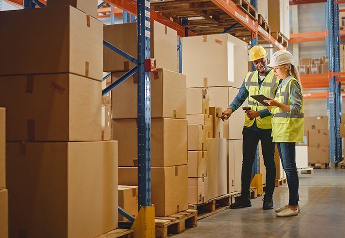 Wo workers checking boxes in a large warehouse wearing safety gear