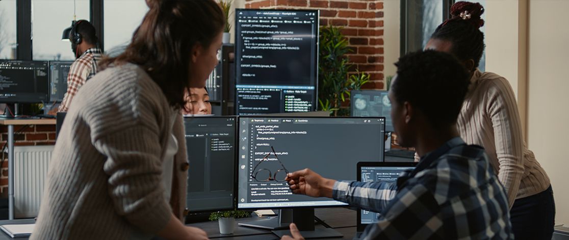  A seated man gestures towards his computer screen while two standing women look at his screen.