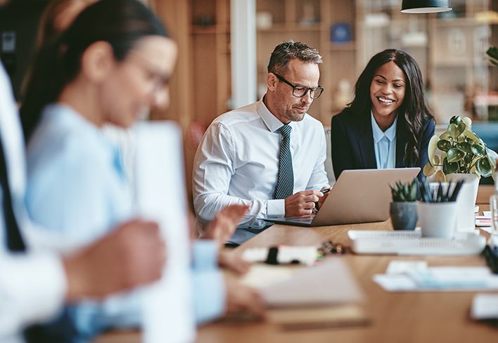 Man dressed in a tie and wearing glasses and woman at a conference table looking at the man's laptop.